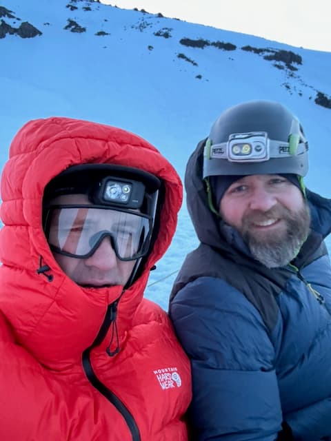 Pre-dawn selfie of Kevin in red puffy with headlamp and Graham in blue puffy with Petzl headlamp on his helmet, both smiling on the snow